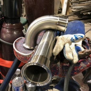 May include: A stainless steel pipe fitting with a 90-degree bend and a threaded end. The fitting is attached to a larger pipe with a clamp. The fitting is surrounded by a blue and white work glove.