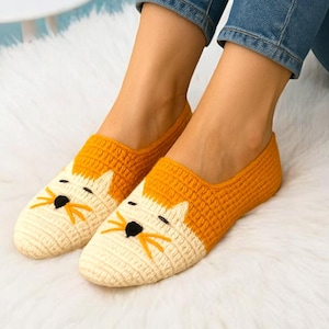 May include: A pair of handmade, crocheted fox slippers in cream and orange. The slippers feature a fox face design with black eyes and nose. The person is wearing blue jeans and sitting on a white fluffy surface.