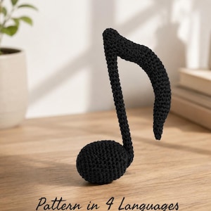 May include: A black crocheted musical note figure with a textured yarn surface. The object is shaped like a standard eighth note with a rounded base and a curved stem. The text "Pattern in 4 Languages" is printed in black at the bottom of the image.