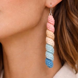 Könnte beinhalten: Nahaufnahme eines farbenfrohen Häkelohrrings. Der Ohrring besteht aus gestapelten, abgerundeten Segmenten in Rosa, Gelb, Blau und Hellblau. Der Text "Pattern in 4 Languages" befindet sich unten.