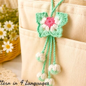 May include: A beige canvas tote bag with a crochet butterfly charm attached to the handle. The charm features a pink center, white petals, and light green edges, with four dangling strings ending in small white and light green crochet spheres.