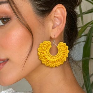 Puede incluir: Un par de pendientes de ganchillo en color amarillo brillante con forma semicircular, que presentan un diseño texturizado en forma de abanico con puntadas intrincadas. Los pendientes se sujetan con un gancho de metal plateado.
