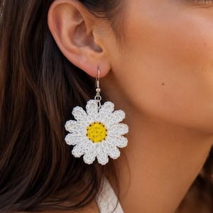 May include: A crochet earring featuring a white daisy flower with a bright yellow center. The flower is attached to a silver-toned metal hook. The design consists of twelve white petals surrounding a textured yellow circular core, creating a floral shape.