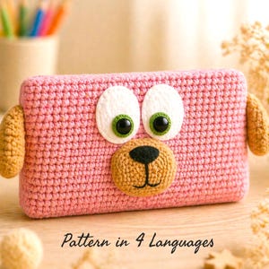 May include: A pink crocheted dog-shaped case with large white eyes, green eye rims, and a tan nose. The case has tan ears on each side. The text "Pattern in 4 Languages" is at the bottom of the image.