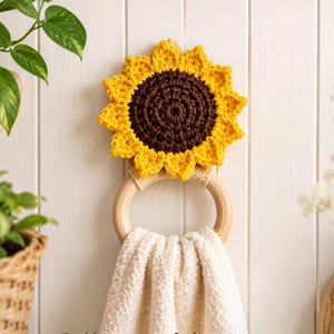 Op de afbeelding: Een gehaakte zonnebloem handdoekhouder met een donkerbruin centrum en felgele bloemblaadjes bevestigd aan een gladde houten ring. Een crèmekleurige handdoek met textuur hangt door de ring. De tekst Pattern in 4 Languages is zichtbaar.