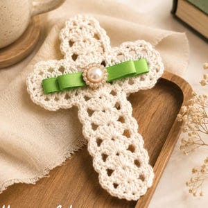 May include: A cream-coloured crocheted cross, adorned with a green ribbon and a pearl-centred embellishment, rests on a wooden tray. The image includes the text "Pattern in 4 Languages" at the bottom.