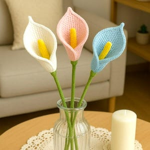 May include: A clear glass vase holds three crocheted calla lilies. The flowers are white, pink, and blue, each with a yellow stamen. The stems are green. The text "Pattern in 4 Languages" is at the bottom of the image.