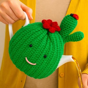 May include: A whimsical green cactus-shaped teapot with a red flower and a ladybug detail, pouring tea into a clear glass. The teapot has a smiling face. The text "Pattern in 4 Languages" is at the bottom.