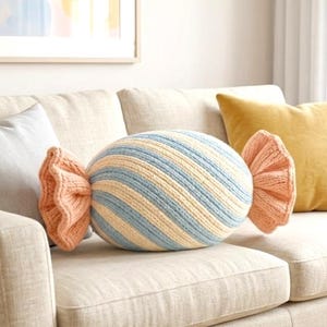 Puede incluir: Un cojín decorativo con forma de caramelo, con rayas azules y crema y extremos con volantes color melocotón, descansa sobre un sofá beige. El texto "Pattern in 4 Languages" es visible. Cojines adicionales en gris y amarillo completan la escena.