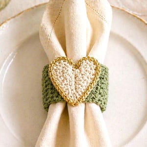 May include: A cream-colored napkin held by a green and gold crochet heart napkin ring, set on a white plate. The image includes a glass, a candle, and text that reads "Pattern in 4 Languages".