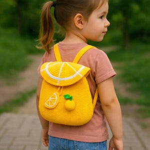 A young girl wearing a bright yellow handmade crochet lemon backpack stands on a tree-lined park path. The backpack features a lemon-slice flap, white crochet details, adjustable shoulder straps, and small lemon-shaped decorations with green leaves. White text at the bottom reads “Pattern in 4 Languages.”