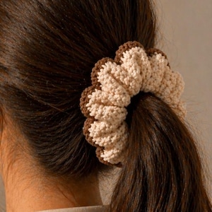May include: A close-up of a ponytail secured with a handmade crochet scrunchie. The scrunchie features a scalloped design with alternating beige and brown sections. The text "Pattern in 4 Languages" is at the bottom.