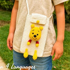 Könnte beinhalten: Eine cremefarbene gehäkelte Umhängetasche für das Telefon mit einem gelben Winnie-Puuh-Charakter. Die Tasche hat einen Klappenverschluss und einen langen Riemen. Der Text "Pattern in 4 Languages" ist unten im Bild zu sehen.