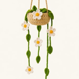May include: A crocheted hanging basket featuring daisy and leaf details. The basket is beige, with green leaves and white daisies with yellow centres. The hanging cords are a natural beige colour. The text "Pattern in 4 Languages" is at the bottom.