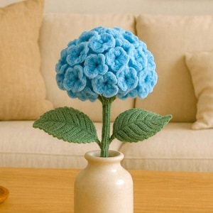 Puede incluir: Una flor de hortensia azul tejida a ganchillo en un jarrón crema, con hojas y tallo verdes. El texto "Pattern in 4 Languages" está en la parte inferior de la imagen. Objeto decorativo.
