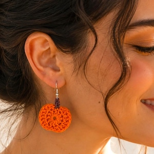 Puede incluir: Un pendiente de calabaza de ganchillo con un cuerpo circular de color naranja vibrante y un pequeño tallo marrón oscuro en la parte superior. La calabaza está unida a un gancho de metal plateado. El texto "Pattern in 4 Languages" es visible.