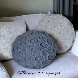 May include: Two round, crocheted moon-shaped cushions, one grey and one beige, with crater detailing. They rest on a white sofa, with a textured cushion to the left. The text "Pattern in 4 Languages" is visible at the bottom.