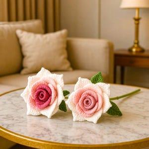 May include: Two handmade crochet roses with pink and white petals and green leaves. The center of each rose is a darker shade of pink. The roses are displayed on a marble-topped table.