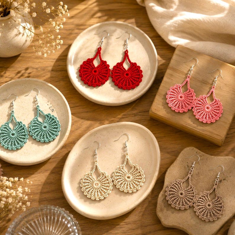May include: A collection of handmade crochet earrings in various colors, including red, teal, pink, beige, and brown. The earrings feature a circular, openwork design with a scalloped edge and silver-tone ear wires. The earrings are displayed on small plates and wooden surfaces.