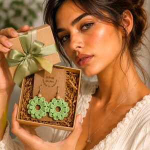 May include: A woman holds a gift box with a pair of green crochet flower earrings. The earrings have wooden beads and silver hooks. The box is brown with a green ribbon and bow. The earrings are displayed on a brown card with text.