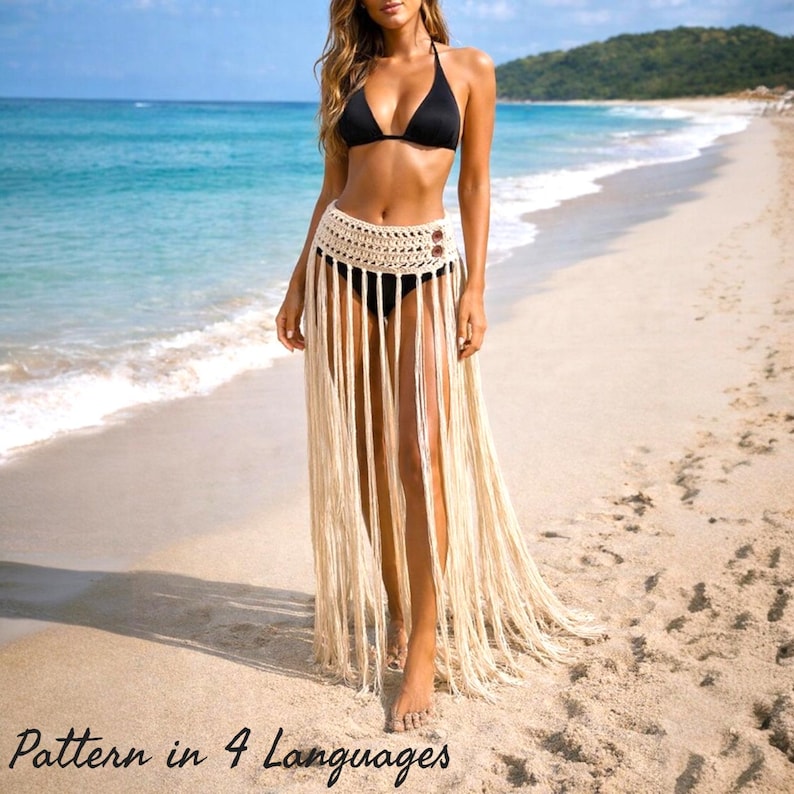 Woman wearing a handmade crochet fringe beach skirt over a bikini, walking on a sandy beach. This crochet skirt features a high waist design with long flowing fringe and boho style details, creating a lightweight and elegant summer look. Perfect as a beach cover up or festival outfit. This crochet skirt pattern PDF includes step by step instructions and size tips for customization, available in four languages.