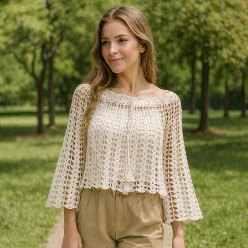 May include: Cream-colored crochet top with bell sleeves and a square neckline. The top features a delicate open weave design and a drawstring detail at the neckline. The garment is paired with tan-colored pants, creating a soft, neutral palette.