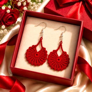 May include: A pair of red crochet earrings with silver-colored hooks, presented in a red gift box. The earrings have a circular design with a scalloped edge. The box is surrounded by red ribbon and a bouquet of flowers.