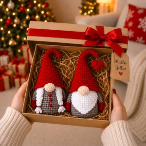 Peut inclure: Un coffret cadeau avec un ruban rouge contient deux gnomes crochet&eacute;s. L'un a une barbe blanche, l'autre des tresses blanches. Les deux ont des chapeaux rouges et des corps gris. La bo&icirc;te porte l'inscription "Mutlu Yillar".