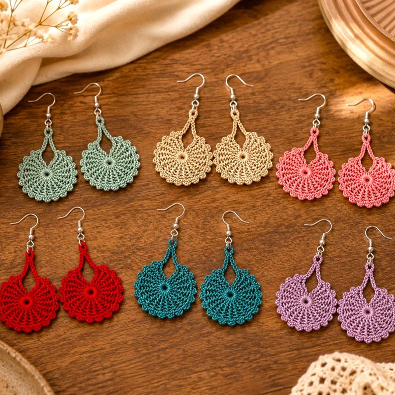 May include: A collection of handmade crochet earrings in various colors, including green, tan, pink, red, teal, and purple. Each pair features a circular design with a central pattern and a decorative top. The earrings are displayed on a wooden surface.