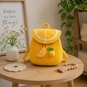 May include: A bright yellow crocheted backpack featuring a flap designed to resemble a sliced lemon with white detailing. The bag includes a small lemon slice applique and a dangling lemon charm with a green leaf, all crafted from yarn.