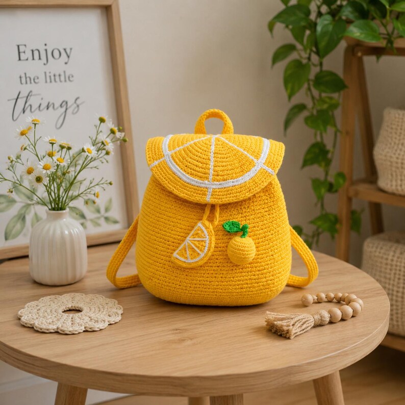 May include: A bright yellow crocheted backpack featuring a flap designed to resemble a sliced lemon with white detailing. The bag includes a small lemon slice applique and a dangling lemon charm with a green leaf, all crafted from yarn.