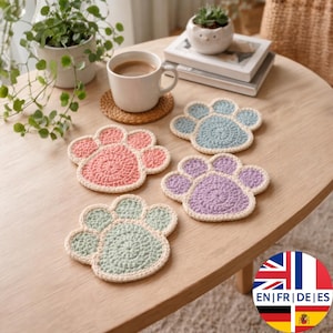 Puede incluir: Cuatro posavasos de ganchillo con forma de pata en rosa, azul, verde y morado, con un borde color crema. Los posavasos están colocados sobre una mesa de madera marrón claro, junto a una taza de café y una pequeña planta.