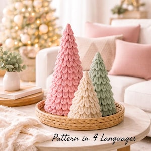 以下が含まれることがあります： ピンク、クリーム、緑色の3つの装飾的なニットクリスマスツリーが、丸いテーブルの織りトレイに置かれています。木は質感のあるレイヤーデザインです。「Pattern in 4 Languages」というテキストが画像の下部にあります。
