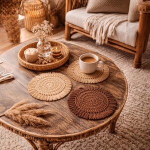 May include: A round, wooden coffee table with four woven coasters, a white mug of coffee, and a decorative tray. The coasters are in shades of beige and brown. A vase of dried flowers and a candle sit on the tray.