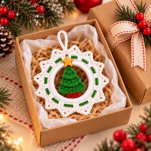May include: A round, white crocheted ornament featuring a green crocheted Christmas tree in the center, topped with a small yellow star. The ornament has a scalloped edge with green accents and a loop at the top, presented inside a brown gift box.