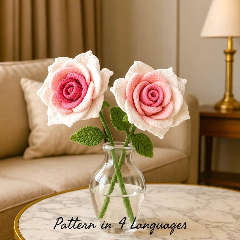 Two handmade crochet roses in soft pink and cream tones are arranged in a small clear glass vase filled with water, placed on a round marble coffee table with a gold rim. The textured petals and green leaves are carefully crafted, giving a realistic floral look. In the background, a cozy beige sofa and a warm-toned lamp create a calm, elegant living room atmosphere with soft natural lighting and a neutral aesthetic.