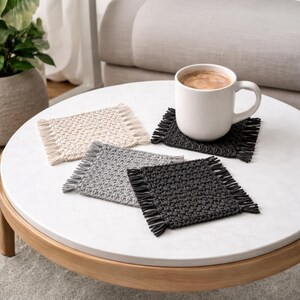 Op de afbeelding: Vier vierkante gehaakte onderzetters in cr&egrave;me, grijs en zwart met franjes worden getoond op een witte tafel. Een witte mok gevuld met koffie staat op een van de zwarte onderzetters. De onderzetters zijn een decoratief woonaccessoire.
