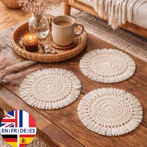 May include: Three cream-colored, round crochet coasters with fringed edges are displayed on a wooden table. The coasters have a spiral design in the center. A mug of coffee, a candle, and a decorative tray are also on the table.