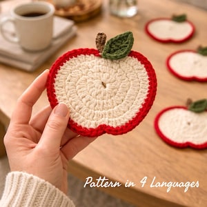 Pode incluir: Aparadores de copos em forma de maçã de crochê com borda vermelha, centro creme e detalhe de folha verde. O texto "Pattern in 4 Languages" é visível. Estes aparadores de copos feitos à mão protegem as superfícies.