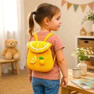 May include: A bright yellow crocheted backpack shaped like a citrus fruit. The flap features a white segment design resembling an orange slice. A small crocheted orange slice charm and a round orange fruit charm hang from the front drawstring closure.