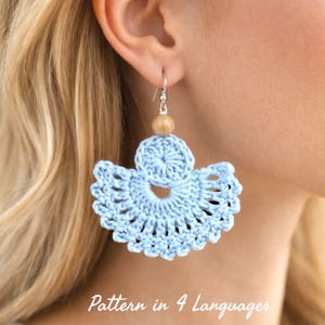 May include: A light blue, crocheted earring with a fan-like design. The earring has a wooden bead and silver-colored hook. The text "Pattern in 4 Languages" is at the bottom of the image. The earring is worn by a person with blonde hair.