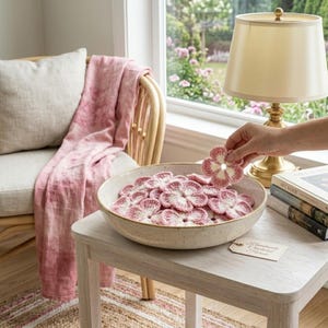 May include: A bowl filled with handmade, pink and white crocheted flower coasters. A hand holds one coaster above the bowl. A lamp and books sit on a light wood table next to a chair with a pink blanket.
