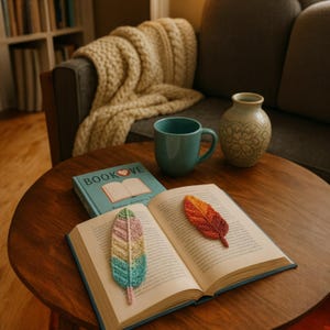 May include: A cozy scene with a wooden table featuring a book with a "BOOK LOVE" cover, a teal mug, and two crocheted feather bookmarks. One bookmark is multi-colored, the other is orange and red. A beige throw blanket and a vase are in the background.