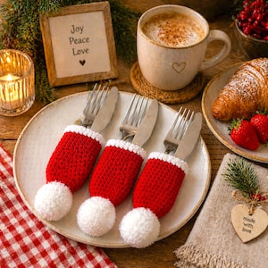 Peut inclure: Une table de No&euml;l festive avec trois porte-ustensiles rouges et blancs au crochet avec des pompons. Une assiette contient les supports avec des fourchettes et des couteaux. Une tasse de caf&eacute;, un croissant et des fraises sont &eacute;galement pr&eacute;sents.