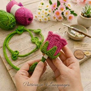 May include: A handmade crochet flower-shaped charger cover in pink and green. The charger cover is held in hands, with a green stem and leaves. The image also shows green and pink yarn balls, and the text "Pattern in 4 Languages".