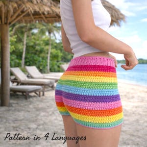 May include: Rainbow-striped crochet shorts with a pink waistband. The shorts are made of colorful yarn in a horizontal striped pattern. The text "Pattern in 4 Languages" is at the bottom of the image. The shorts are worn on a beach.