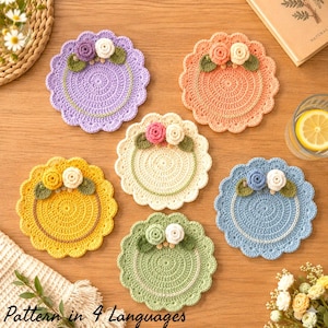 May include: Six round crochet coasters with scalloped edges, each featuring two small crocheted flowers and green leaves. The coasters come in purple, peach, cream, yellow, light blue, and sage green, each with a circular stitched detail.