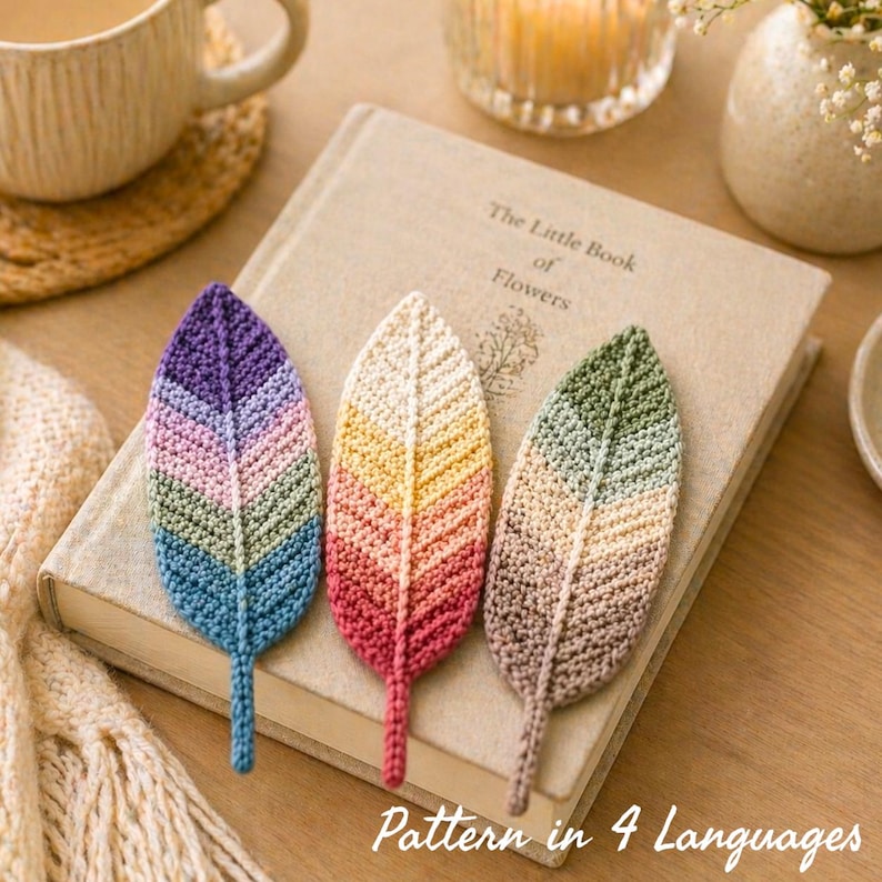 Pastel-toned crochet leaf bookmarks displayed on a closed neutral-colored book, styled in a cozy lifestyle setting with a soft knit blanket, ceramic coffee cup, lit candle, and delicate flowers on a wooden table. The handcrafted bookmarks are placed in the foreground, highlighting their texture and color variations. Warm, natural lighting creates a calm, aesthetic scene perfect for book lovers, emphasizing a handmade, giftable crochet accessory.