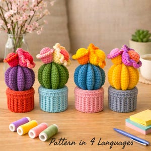 May include: Four crochet cactus ornaments in vibrant colours: purple, green, blue, and yellow, each with flower details. They are displayed on matching crochet pots. The image includes thread spools and the text "Pattern in 4 Languages".