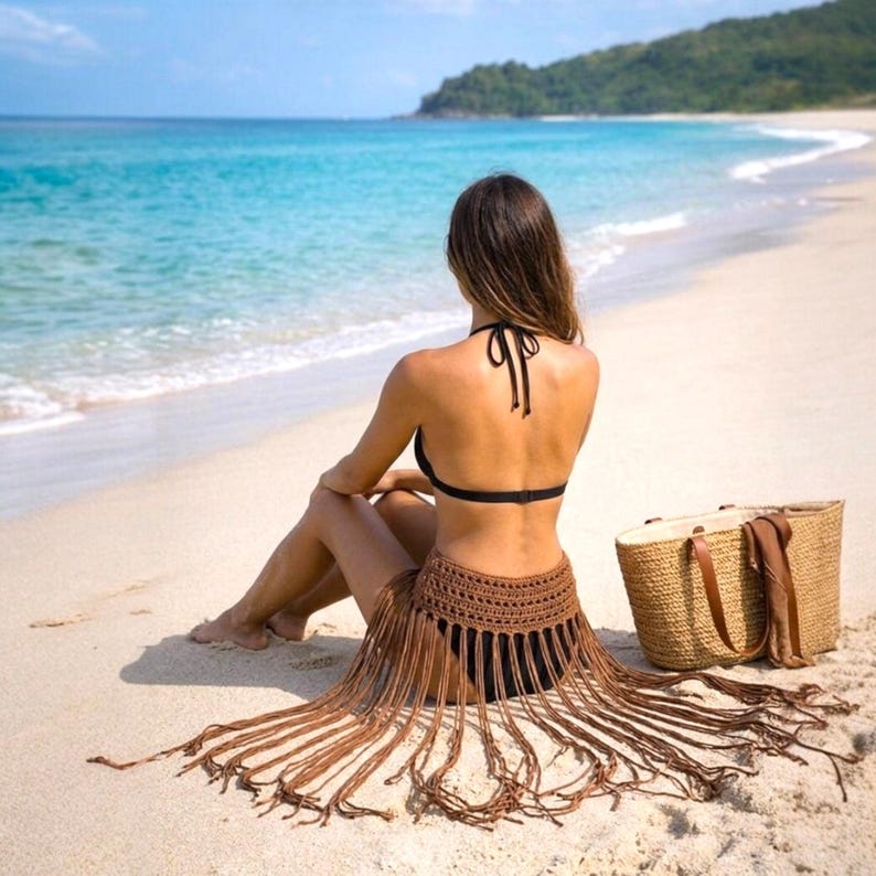 Pode incluir: Uma mulher de biqu&iacute;ni preto e saia de franjas de croch&ecirc; marrom senta-se numa praia arenosa. Uma sacola de palha tecida com al&ccedil;as de couro e um len&ccedil;o marrom est&atilde;o por perto. O mar e uma colina verde est&atilde;o ao fundo.